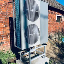 Szara dwuwentylatorowa jednostka zewnętrzna systemu grzewczego marki Technix, zamontowana na stalowej konstrukcji wsporczej przy ścianie z czerwonej cegły, widoczna instalacja elektryczna.