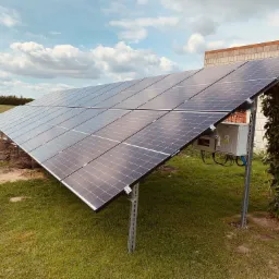 Instalacja paneli słonecznych na konstrukcji wsporczej, widoczna skrzynka elektryczna, w tle pole z turbiną wiatrową i budynek z cegły.