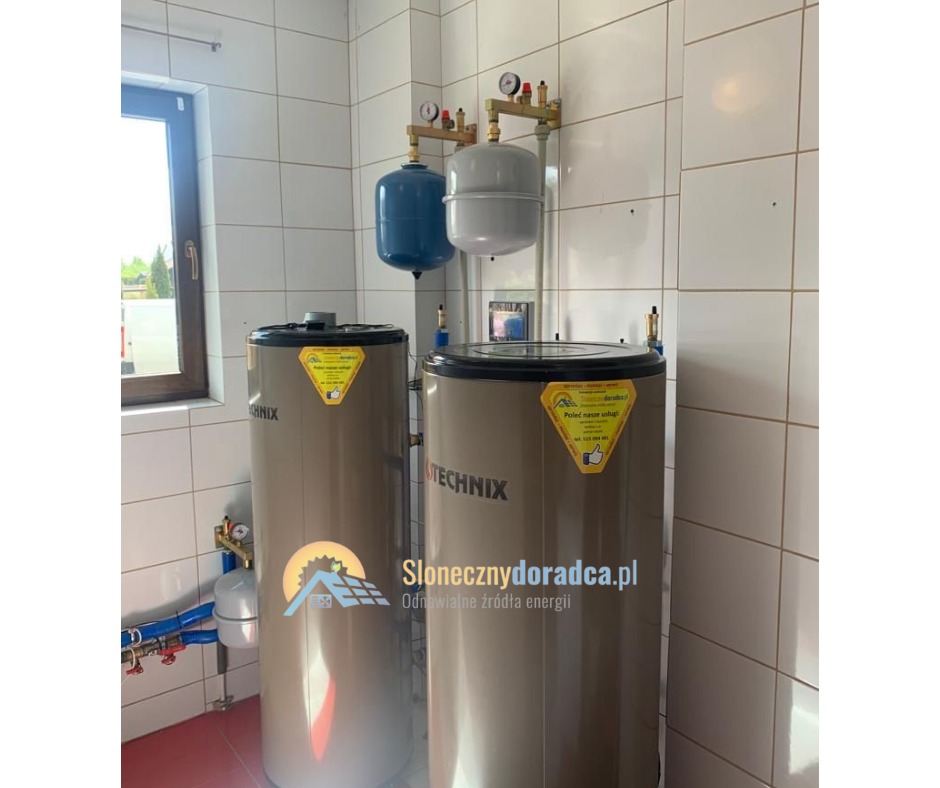 Dwa pionowe zbiorniki buforowe Technix w kotłowni, z niebieskim i szarym naczyniem przeponowym podwieszonymi na ścianie wyłożonej białymi płytkami. Widoczne orurowanie i okno.