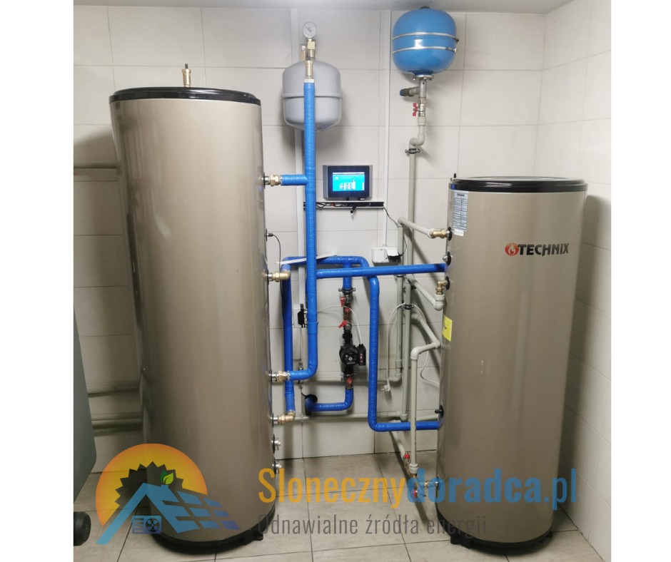 Dwa zbiorniki akumulacyjne ciepła w kotłowni, połączone niebieskimi rurami, z widocznym ekranem sterującym i firmowym logo na jednym ze zbiorników.