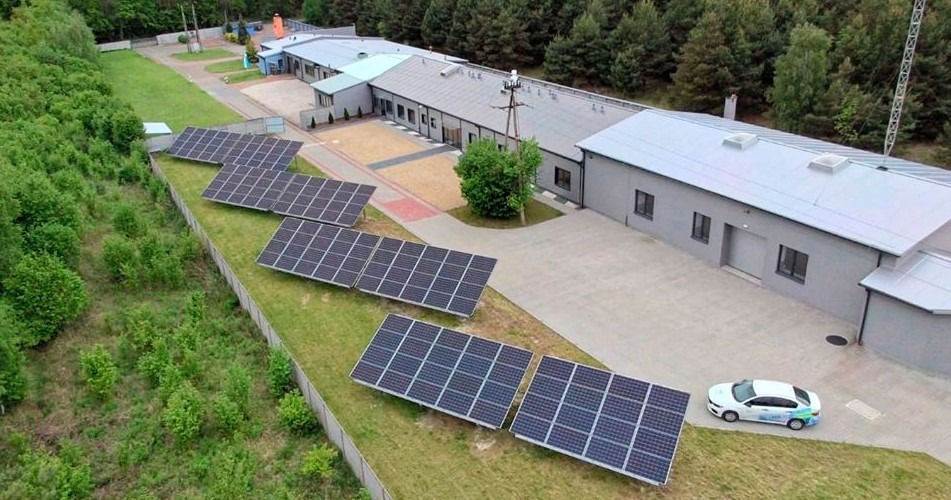 Widok z lotu ptaka na panele słoneczne zainstalowane na trawiastym terenie przylegającym do budynku przemysłowego; w tle las i samochód firmowy.