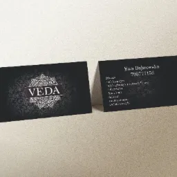 Dwie czarne wizytówki z logo Veda i danymi kontaktowymi Ewy Dąbrowskiej, leżące na beżowym tle. Na jednej wizytówce widoczne logo firmy, na drugiej dane kontaktowe i lista oferowanych masaży.