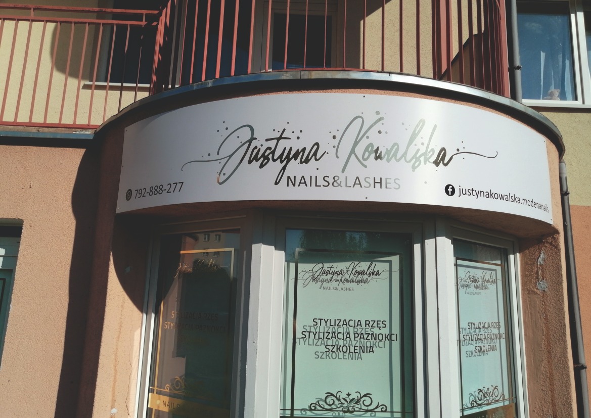 Zewnętrzna tablica informacyjna z nazwą salonu 'Justyna Kowalska Nails & Lashes' nad wejściem, wykonana z białego PCV z ploterowanymi napisami w kolorze złotej lustrzanej folii, widoczny numer...