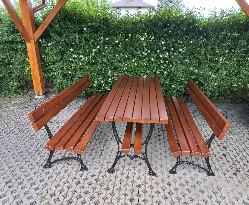 Zestaw mebli ogrodowych: drewniany stół i dwie ławki z oparciami na kutych, czarnych nogach, ustawione na szarej kostce brukowej, w tle zielony żywopłot z białymi kwiatami i fragment drewnianej...