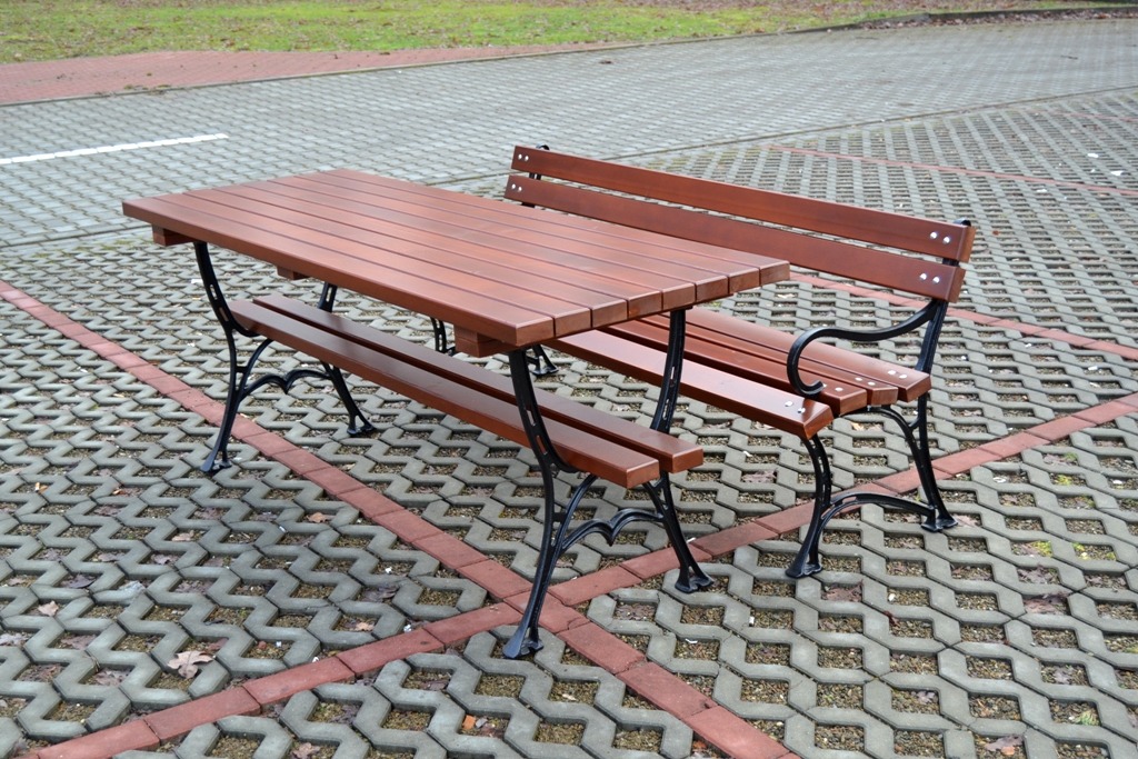 Drewniany stół piknikowy z dwiema ławkami na metalowych nogach, ustawiony na szarej kostce brukowej z bordiurą z czerwonej kostki.
