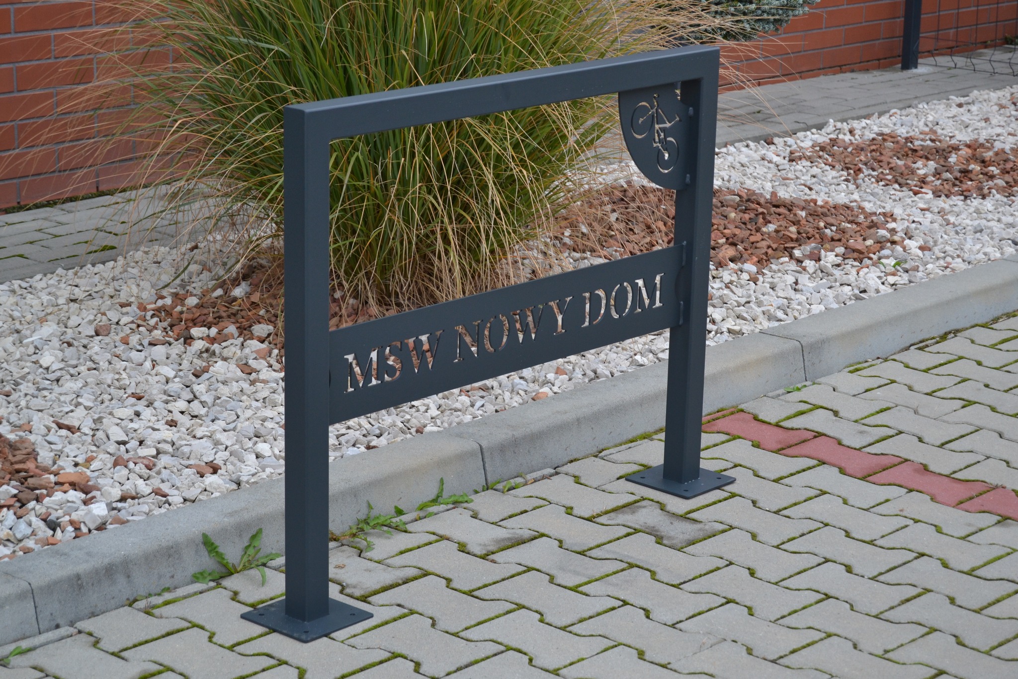 Stalowy stojak rowerowy z wyciętym napisem 'MSW NOWY DOM' oraz symbolem roweru, umieszczony na chodniku z kostki, w tle ozdobne kamienie i trawa.