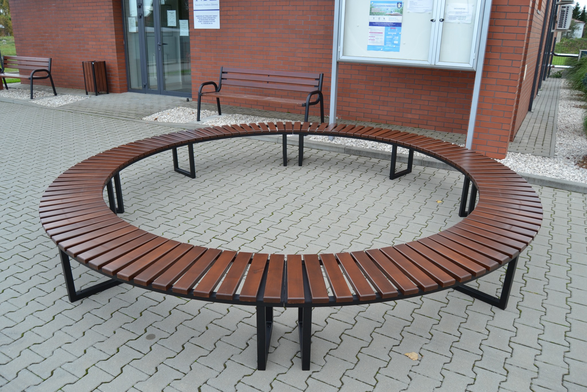 Okrągła ławka z drewnianymi szczebelkami i czarną metalową konstrukcją, umieszczona na brukowanym placu przed budynkiem z czerwonej cegły. W tle widoczne inne ławki i tablica informacyjna.