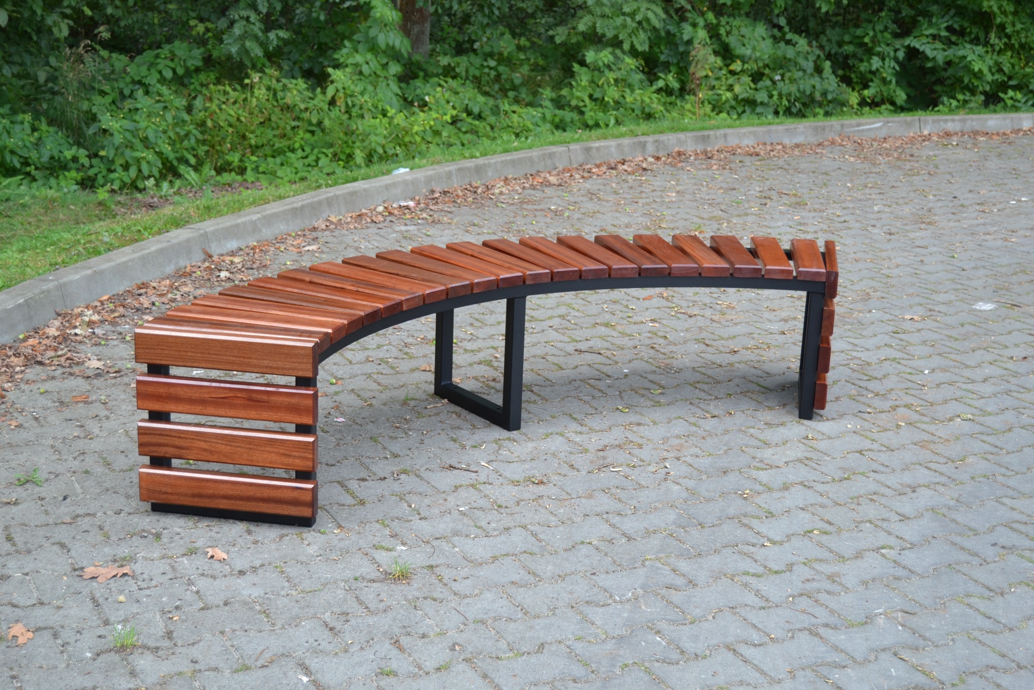 Zakrzywiona ławka parkowa z drewna i metalu na brukowanym placu, z zielenią w tle.