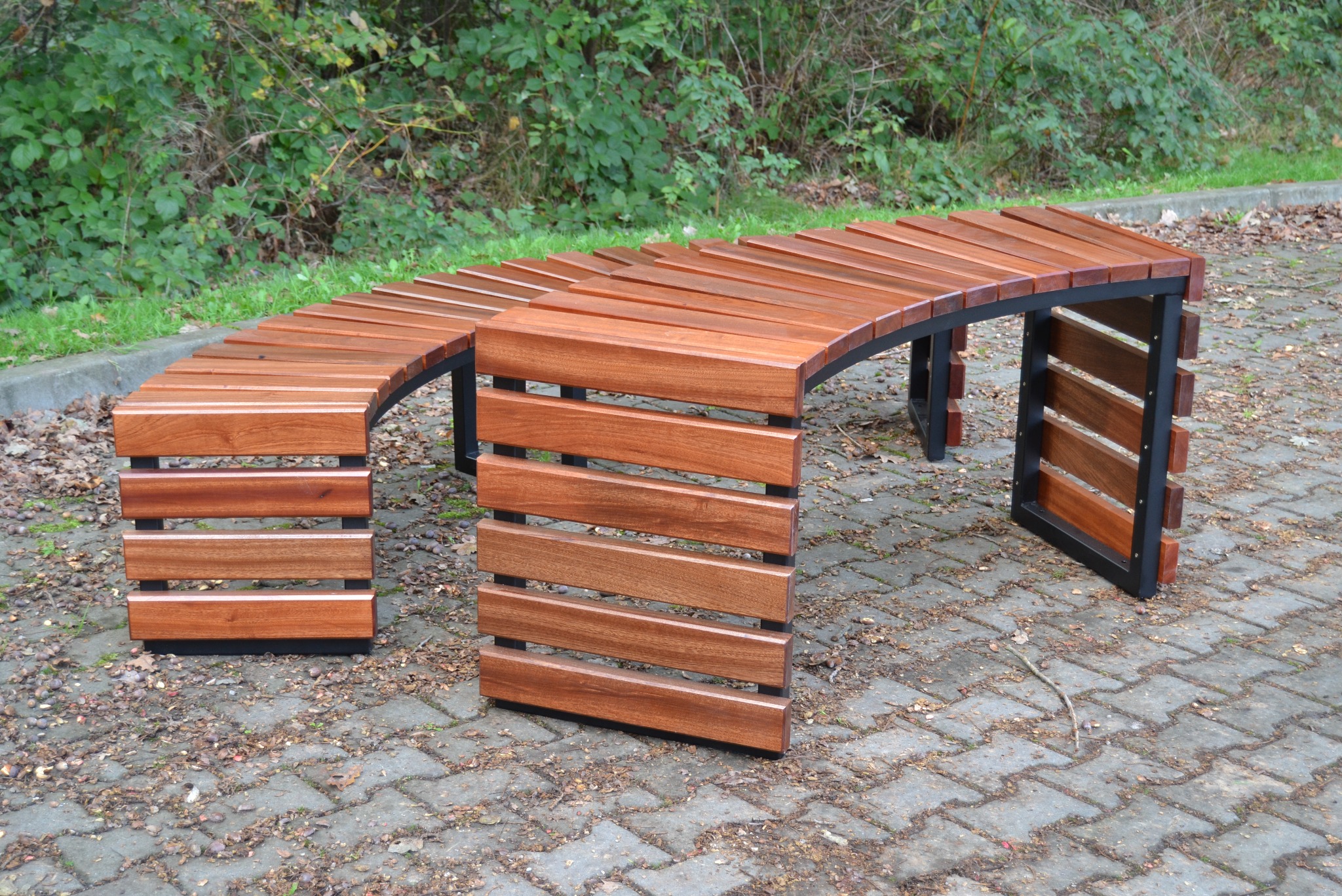 Zakrzywiona ławka parkowa z drewna i metalu, ustawiona na brukowanej nawierzchni, z zielenią w tle.