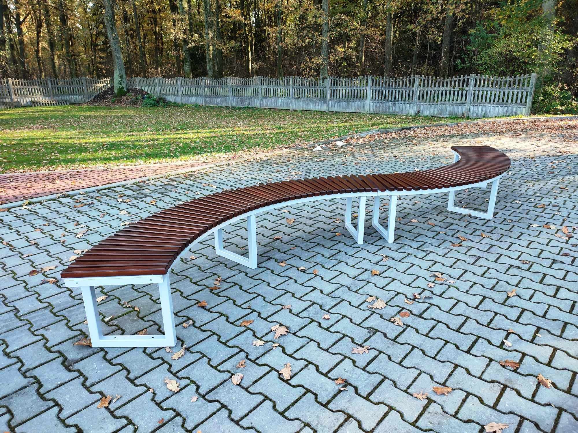 Długa, łukowata ławka parkowa z brązowych listew na białych, metalowych nogach, ustawiona na szarej kostce brukowej z opadłymi liśćmi, w tle trawnik, drzewa i drewniany płot.