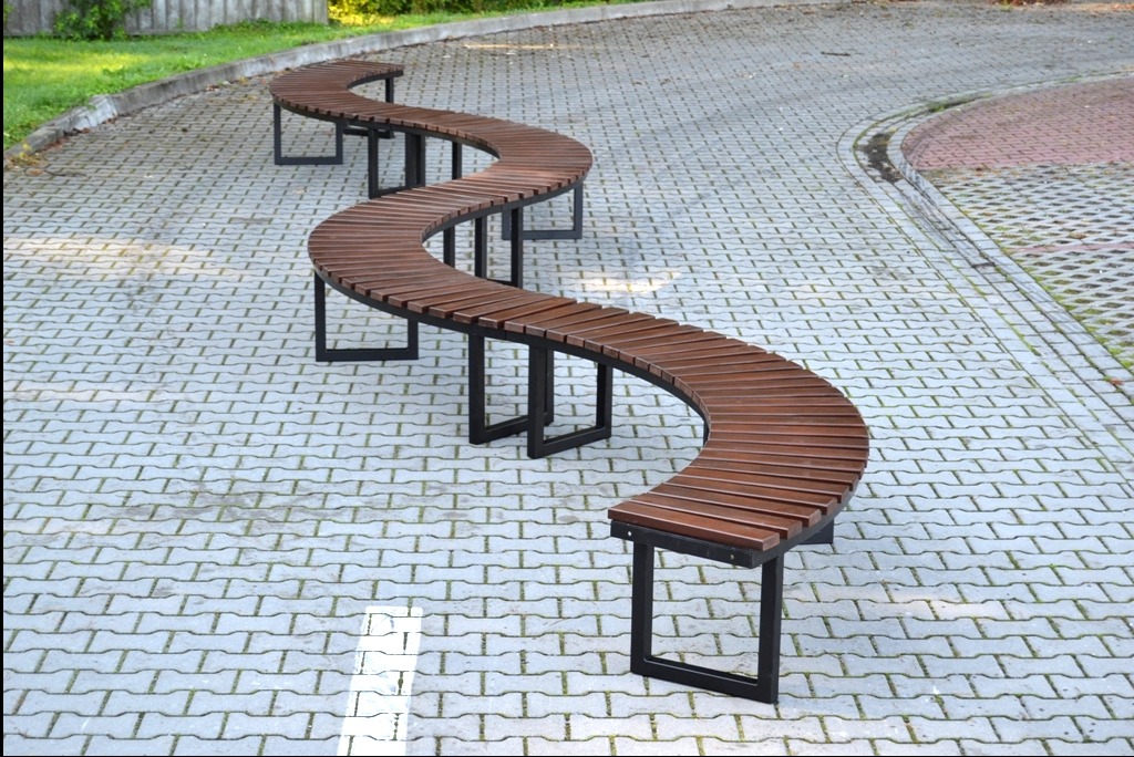 Długa, zakrzywiona ławka parkowa z drewnianym siedziskiem i czarnymi metalowymi nogami, ustawiona na szarej kostce brukowej.