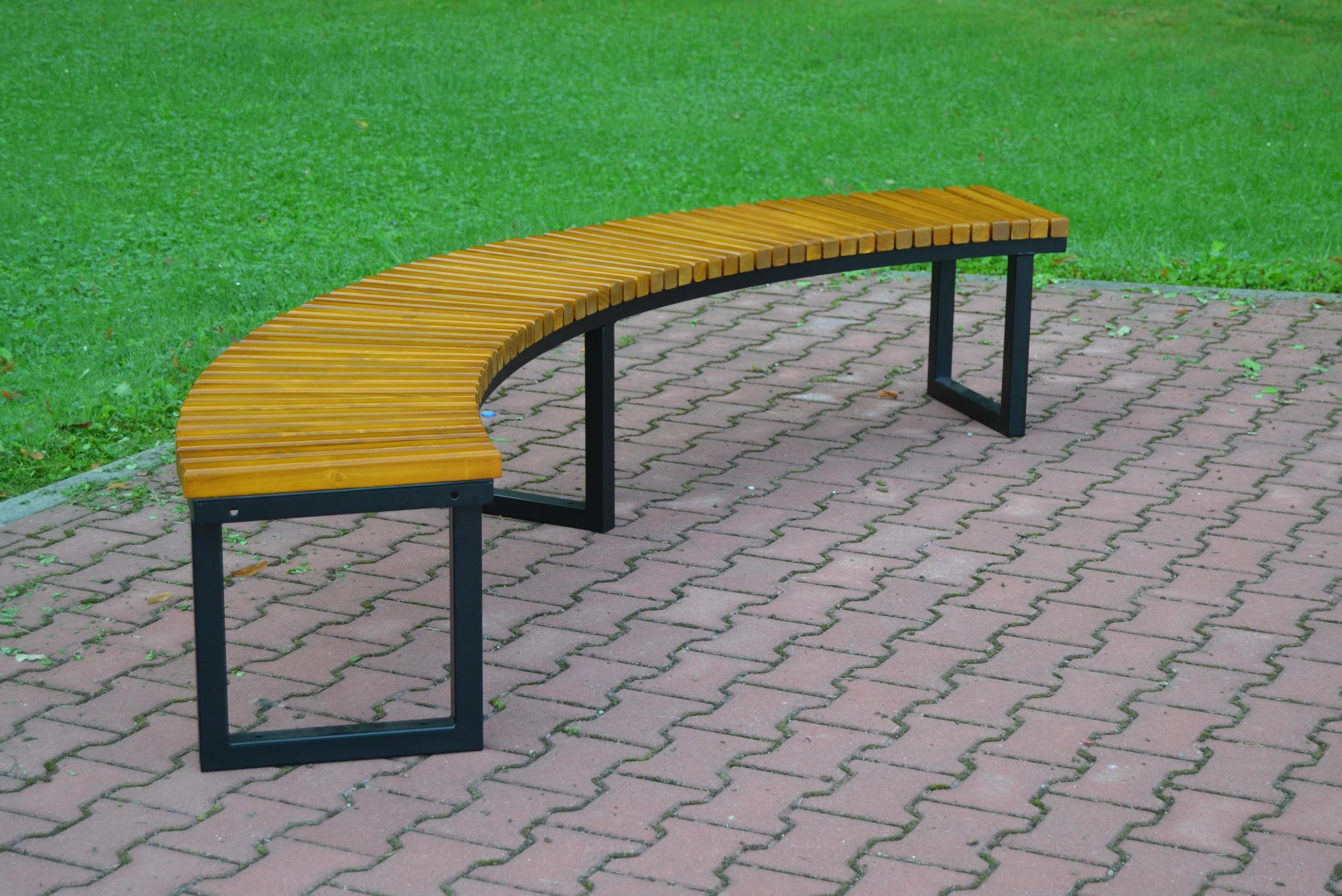 Zakrzywiona ławka parkowa z drewnianym siedziskiem i czarnymi metalowymi nogami, ustawiona na czerwonej kostce brukowej, z zielonym trawnikiem w tle.