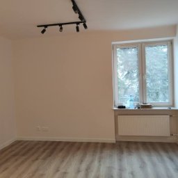 REMONTY I WYKOŃCZENIA KRZYSZTOF PAZIEWSKI - Jasne wnętrze pokoju po odświeżeniu: białe ściany, panelowa podłoga, okno z widokiem na drzewa, czarna listwa oświetleniowa na suficie. Minimalistyczny styl.