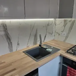 Wykończona kuchnia z marmurowym backsplash, drewnianym blatem i szarymi szafkami. Widoczny zlew, bateria i płyta gazowa. Czysta i nowoczesna estetyka.