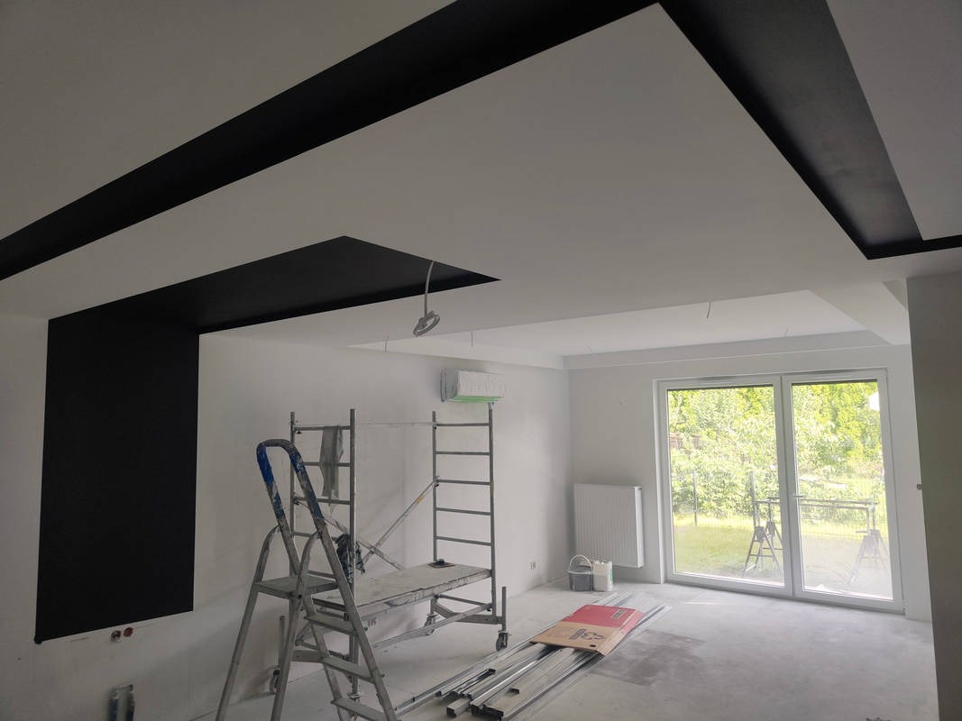 Pokój po malowaniu z czarnymi akcentami na suficie i ścianie. Widoczne rusztowania, okno balkonowe i grzejnik. Podłoga przygotowana do dalszych prac.