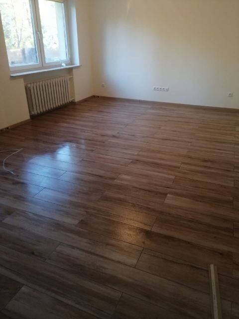 Ukończona podłoga z płytek imitujących drewno w jasnym pokoju z oknem i grzejnikiem. Widoczne fugi i naturalne światło podkreślające strukturę.