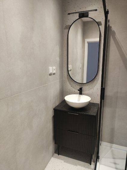 Nowoczesna łazienka z owalnym lustrem w czarnej ramie, umywalką nablatową i ciemną szafką z ryflowanymi frontami. Szare płytki i kabina prysznicowa w tle.