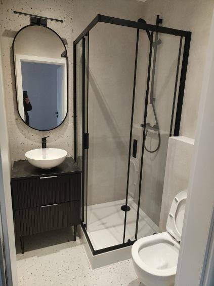 Nowoczesna łazienka z czarną kabiną prysznicową, umywalką na szafce z ryflowanym frontem i owalnym lustrem z podświetleniem. Minimalistyczny design i jasne wykończenie ścian.