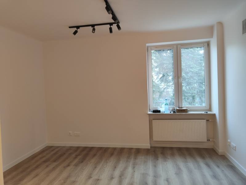 Jasne wnętrze pokoju po odświeżeniu: białe ściany, panelowa podłoga, okno z widokiem na drzewa, czarna listwa oświetleniowa na suficie. Minimalistyczny styl.