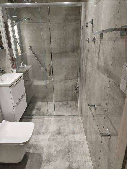 Szara łazienka z kabiną prysznicową, umywalką i toaletą. Widoczne płytki imitujące beton, szklana kabina i chromowane akcesoria. Nowoczesny minimalizm.