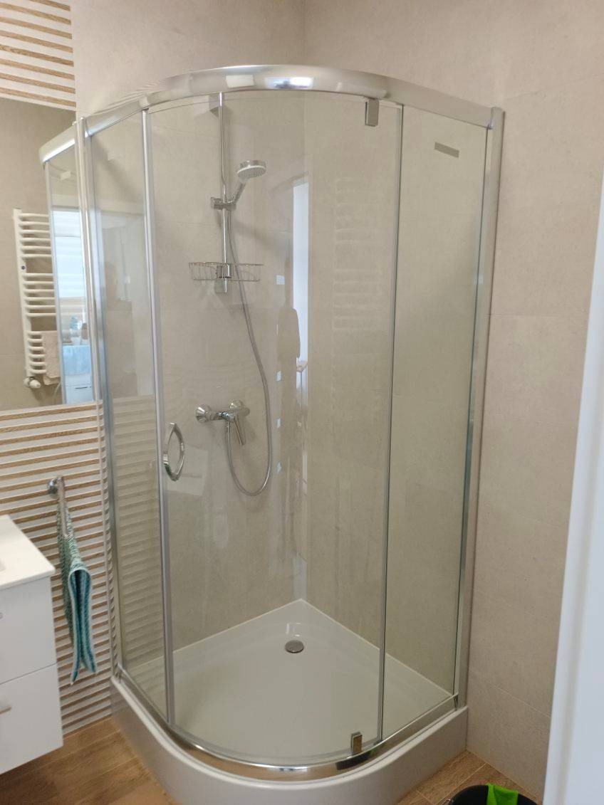Narożna kabina prysznicowa z chromowanymi profilami i szklanymi ściankami, widoczna w wykończonej łazience z beżowymi płytkami i dekoracyjną ścianą.