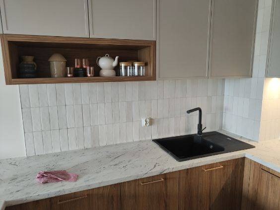 Nowoczesna kuchnia po remoncie: białe płytki na ścianie, marmurowy blat, drewniane szafki z minimalistycznymi uchwytami i czarny zlew.