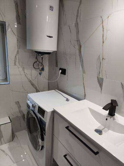 Wykończona łazienka z marmurowymi płytkami, białą szafką z umywalką i czarną baterią. Widoczny bojler nad pralką. Minimalistyczny design, jasne wnętrze.