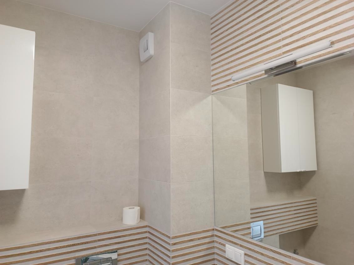 Beżowa łazienka z poziomymi paskami, lustrem i białą szafką. Widoczny fragment toalety i rolka papieru. Oświetlenie górne.