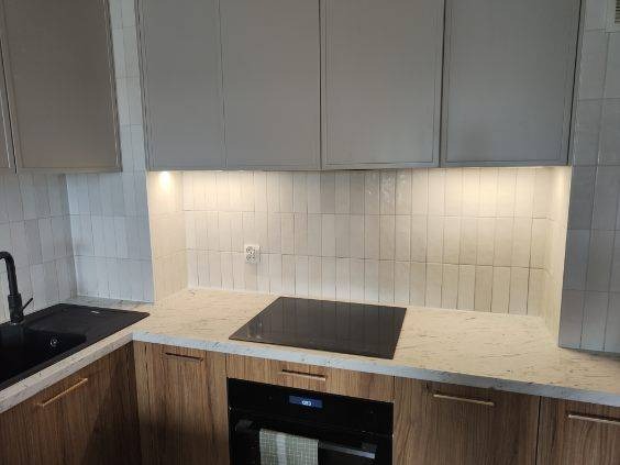 Nowoczesna kuchnia z białymi, pionowo ułożonymi płytkami i drewnianymi szafkami. Czarny zlew i płyta indukcyjna. Oświetlenie LED pod szafkami górnymi.