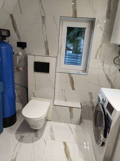 Wykończona łazienka z toaletą, pralką i oknem. Płytki imitujące marmur z brązowymi żyłami pokrywają ściany i podłogę. Widoczny filtr wody.