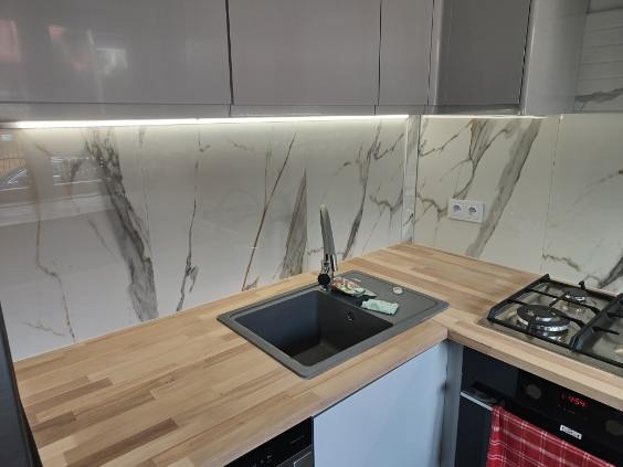 Wykończona kuchnia z marmurowym backsplash, drewnianym blatem i szarymi szafkami. Widoczny zlew, bateria i płyta gazowa. Czysta i nowoczesna estetyka.