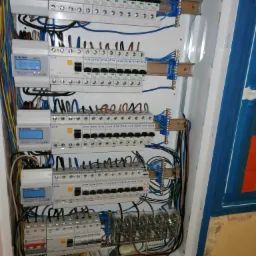 Rozdzielnica elektryczna z zamontowanymi licznikami energii i zabezpieczeniami nadprądowymi, widoczne kolorowe przewody i listwy zaciskowe.