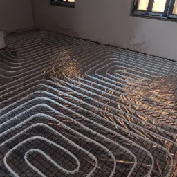 Rozłożona instalacja ogrzewania podłogowego z białych rurek na czarnej siatce montażowej, widoczne okna i przygotowana wylewka w tle.