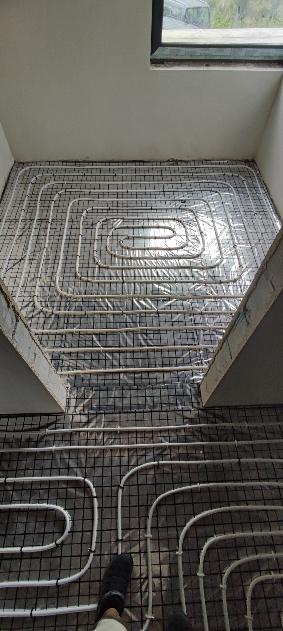 Układanie rur ogrzewania podłogowego na siatce, widoczne białe rury w spiralnym układzie, czarna siatka montażowa i fragment stopy w czarnym bucie.