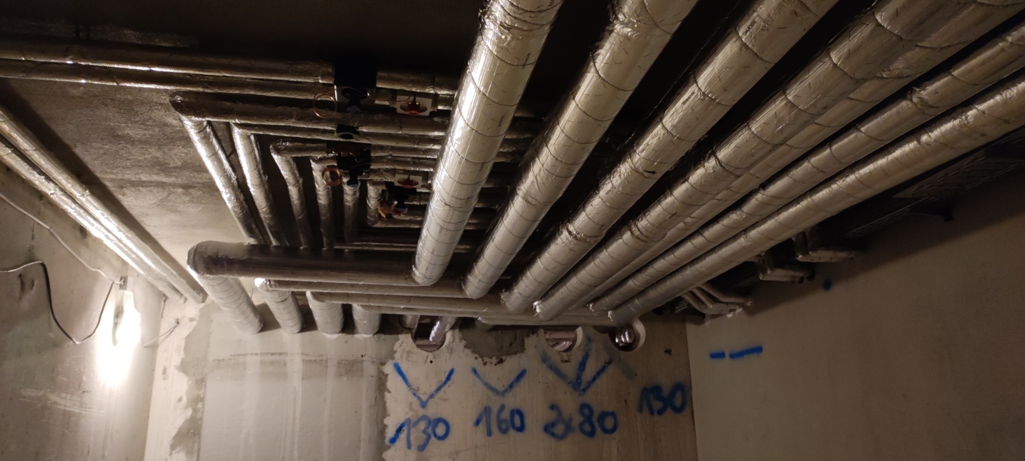 Rzędy zaizolowanych rur pod sufitem w surowym pomieszczeniu, widoczne zawory i niebieskie oznaczenia na ścianie.