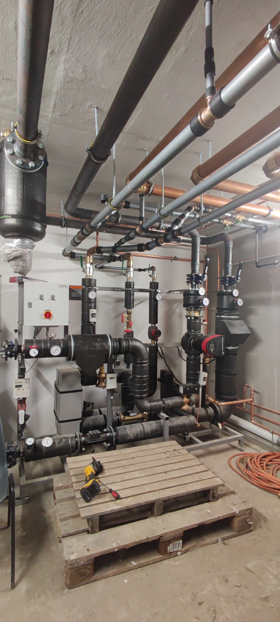 Złożona instalacja grzewcza z rur miedzianych, stalowych i plastikowych, zaworami, manometrami i czujnikami, narzędzia na palecie pod instalacją, surowy betonowy sufit.