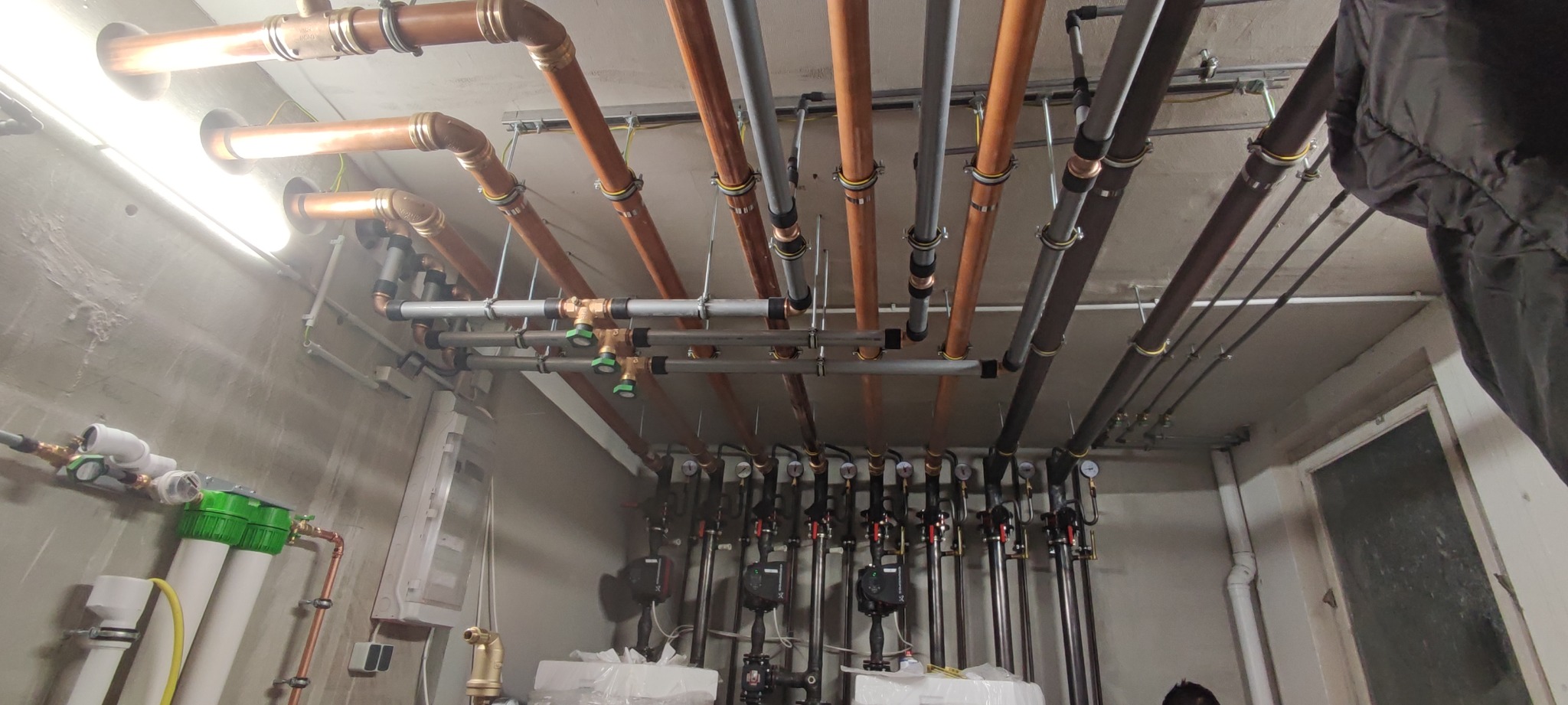 Skomplikowany system rur miedzianych i stalowych pod sufitem, z widocznymi zaworami, manometrami i pompami obiegowymi, zamontowany w kotłowni lub pomieszczeniu technicznym.