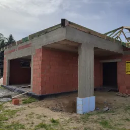 Wznoszenie konstrukcji budynku z czerwonej cegły, widoczne betonowe słupy i zbrojenie dachu, ocieplenie fundamentów niebieskim styropianem.