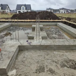 Zalany wodą wykop pod fundamenty domu jednorodzinnego z wystającymi prętami zbrojeniowymi, w tle hałda ziemi i zabudowa osiedlowa.