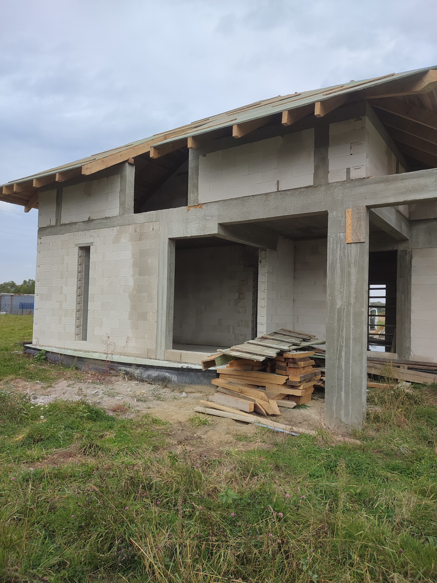 Surowy stan domu jednorodzinnego z betonu komórkowego, widoczne fundamenty, ściany i zarys dachu, składowane deski przy filarze.