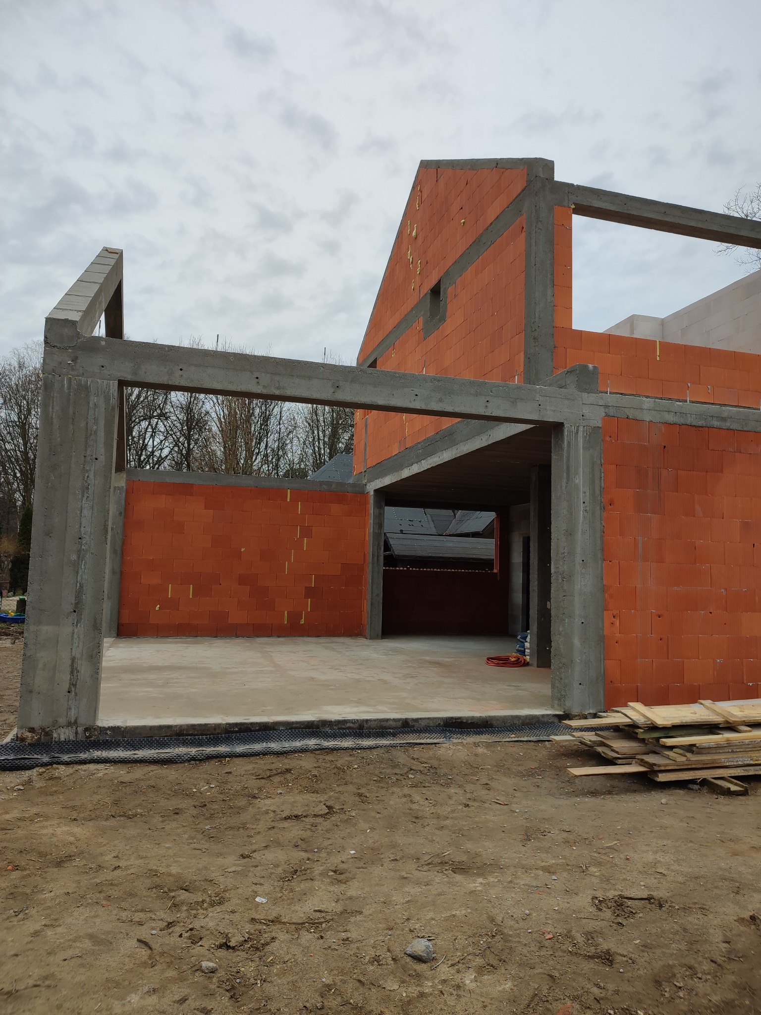 Surowy stan domu murowanego z betonu i pustaków ceramicznych, widoczne zbrojenia, fundamenty zabezpieczone folią, w tle drzewa i pochmurne niebo.