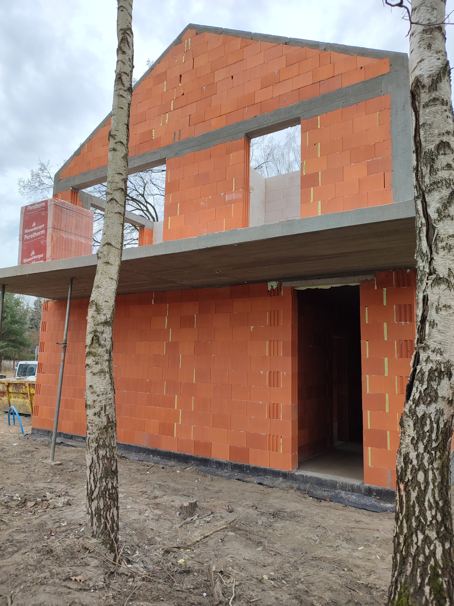 Surowy stan dwukondygnacyjnego budynku w trakcie budowy, wzniesionego z czerwonej cegły Porotherm, z widocznymi otworami okiennymi i betonowym stropem, na pierwszym planie brzoza.