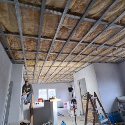 FRB Marcin Zawistowski - Montaż izolacji z wełny mineralnej na poddaszu z widoczną konstrukcją metalową, pracownik na podeście, materiały budowlane i narzędzia w pomieszczeniu.