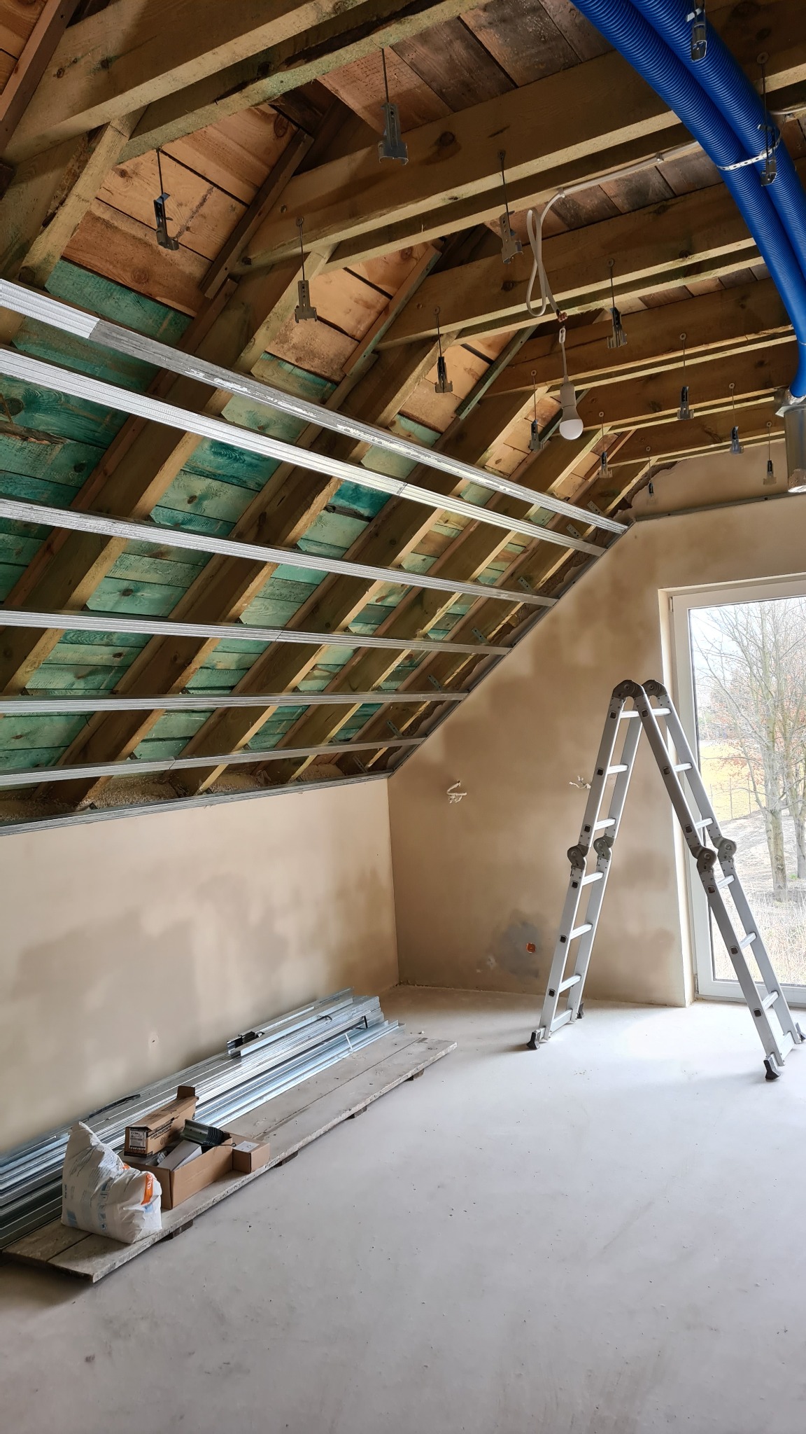 Poddasze w trakcie przygotowania do montażu płyt gipsowo-kartonowych, widoczne metalowe profile sufitowe, drewniana konstrukcja dachu z instalacją, drabina przy oknie i materiały budowlane...