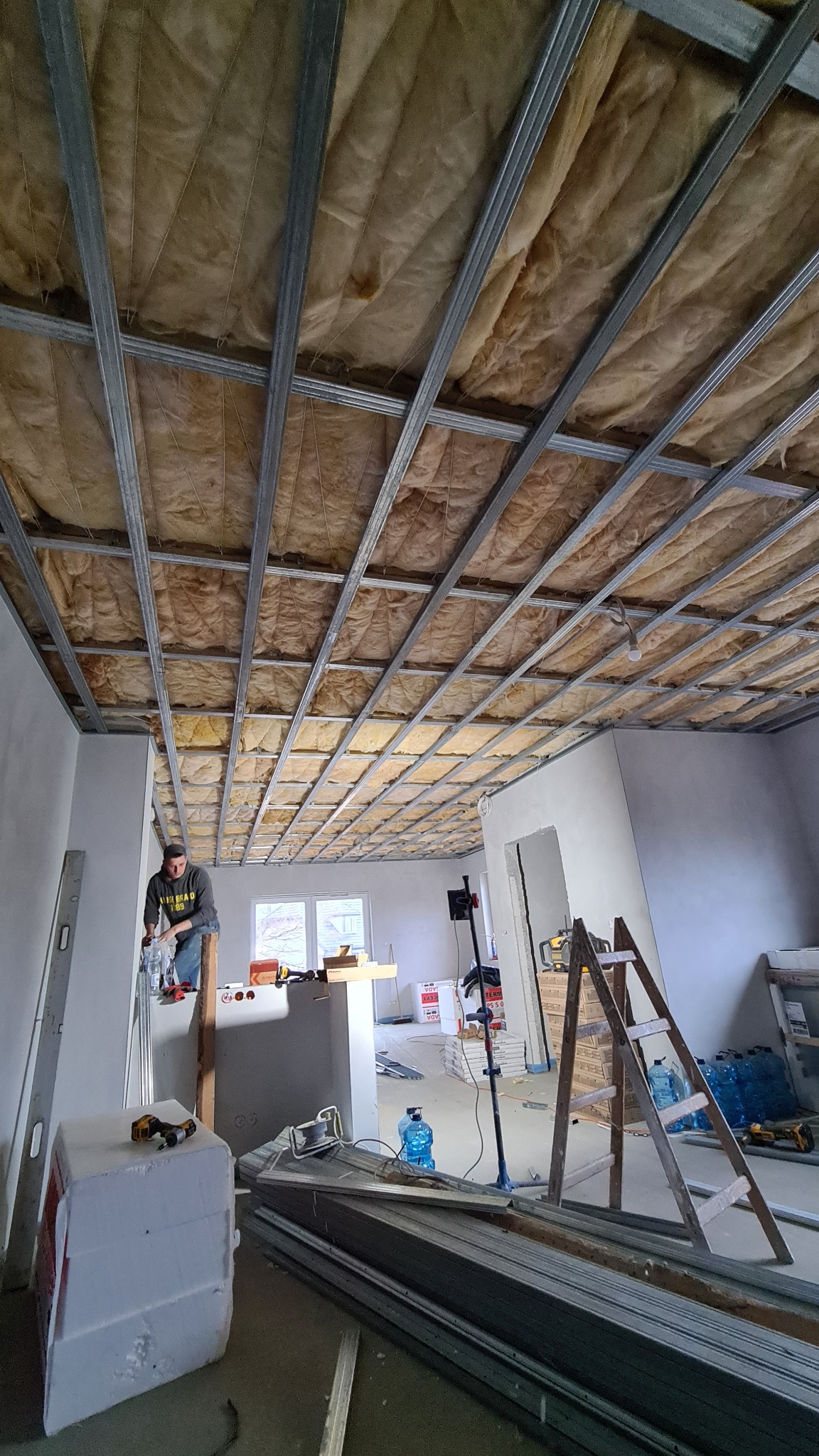 Montaż izolacji z wełny mineralnej na poddaszu z widoczną konstrukcją metalową, pracownik na podeście, materiały budowlane i narzędzia w pomieszczeniu.