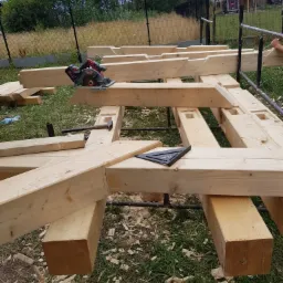 Konstrukcja z jasnego drewna na rusztowaniu, widoczna piła tarczowa, młotek i kątownik, w tle trawa i ogrodzenie.