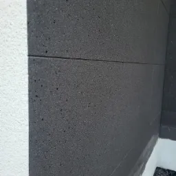 Fragment ściany budynku z widocznym ociepleniem z grafitowego styropianu, obok fragment otynkowanej ściany pomalowanej na biało.