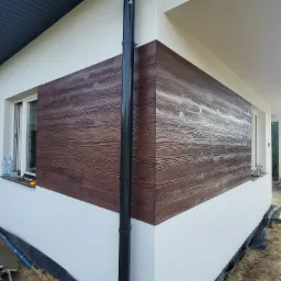 Wykończona elewacja domu jednorodzinnego z brązową okładziną imitującą drewno, połączona z białym tynkiem, widoczna rura spustowa i teren wokół w trakcie prac ziemnych.