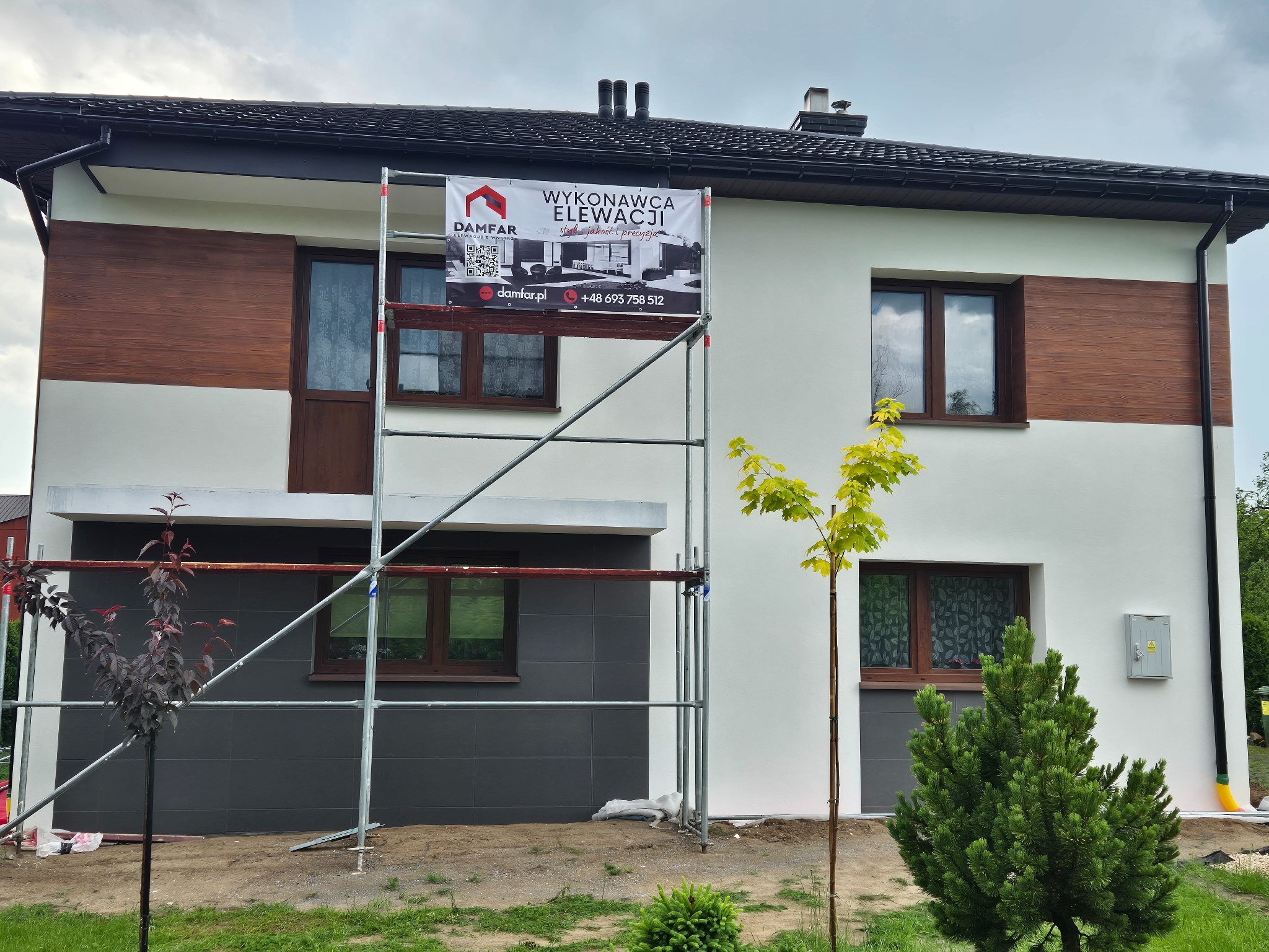 Dwukondygnacyjny dom z elewacją w odcieniach bieli, szarości i drewna. Na rusztowaniu baner reklamowy wykonawcy elewacji. Widoczne drzewka i krzewy.