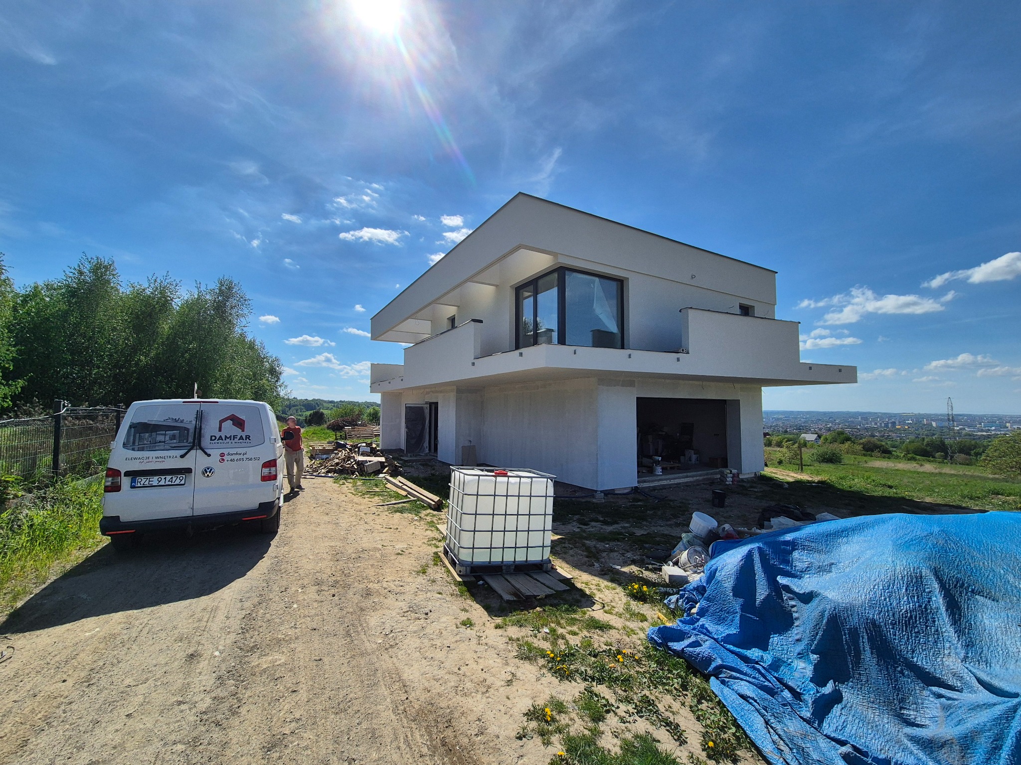 Nowoczesny, dwukondygnacyjny dom w trakcie budowy, widoczny z zewnątrz, z białymi ścianami i dużymi oknami. Na pierwszym planie biały zbiornik i auto dostawcze.